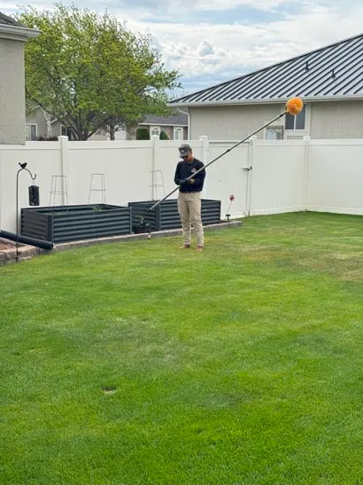 Bliss technician reaching roof eaves from a backyard lawn, residential pest control in Salt Lake City
