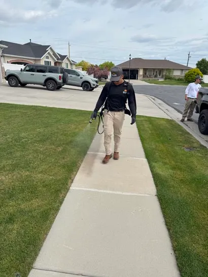 Bliss technician spraying a lawn edge beside a sidewalk, exterior pest control in Salt Lake City
